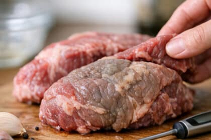 Close-up of raw beef on a wooden cutting board with spices and a meat thermometer inserted.