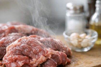 Fresh raw beef steaks on a wooden cutting board with garlic cloves and seasoning in the background.