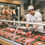 Soğutmalı reyonda kırmızı et düzenleyen kasap ve arka planda cam kapılı dry aged dolabı